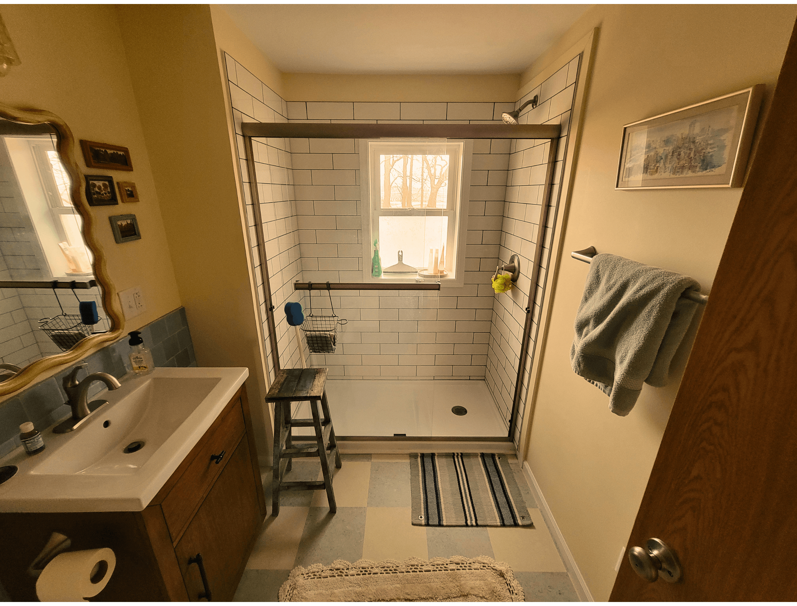 Bathroom with white subway tile shower, window, wooden vanity, and a small wooden stool.