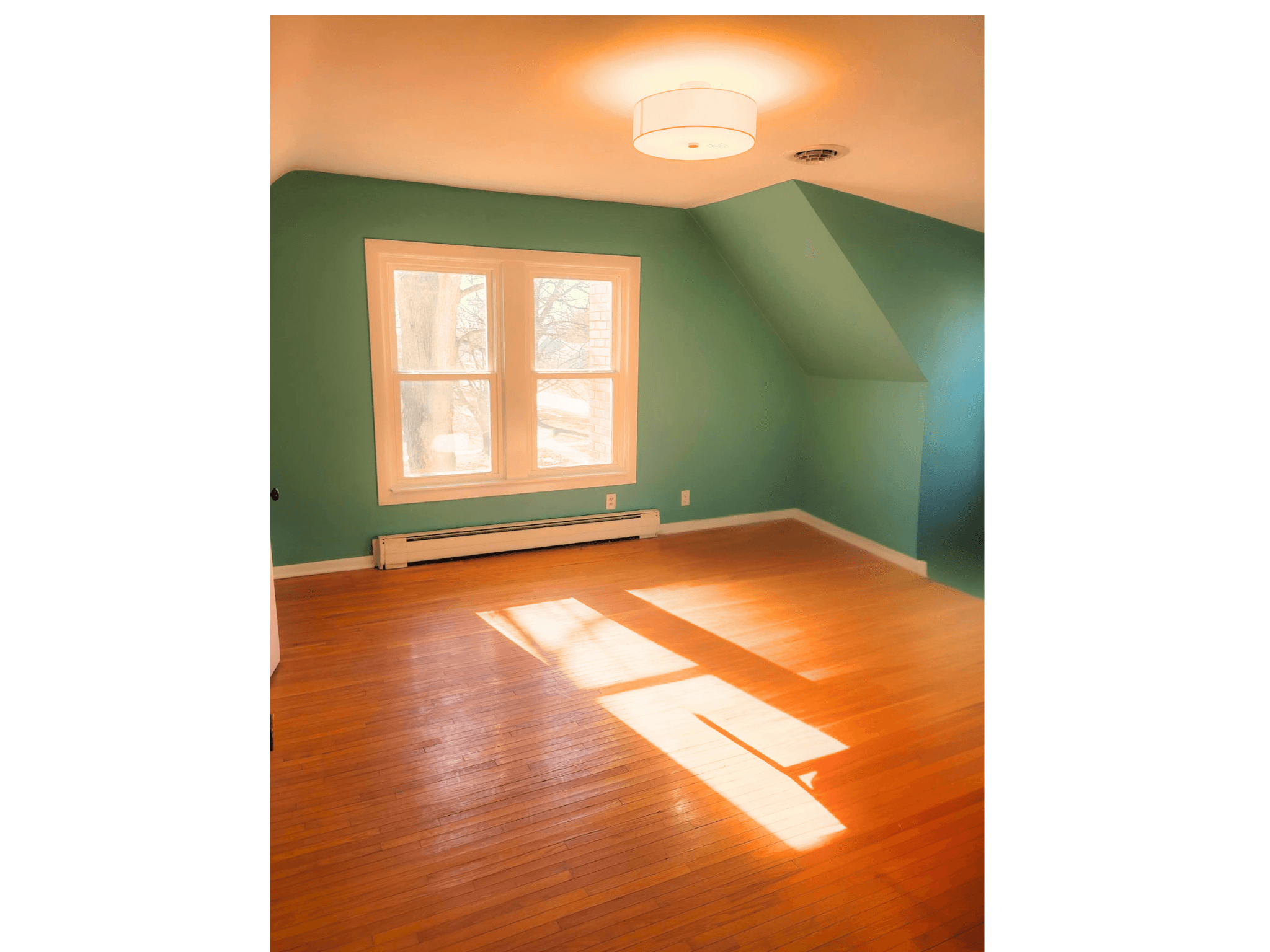 Empty room with teal walls, hardwood floors, and bright sunlight streaming through two white windows.