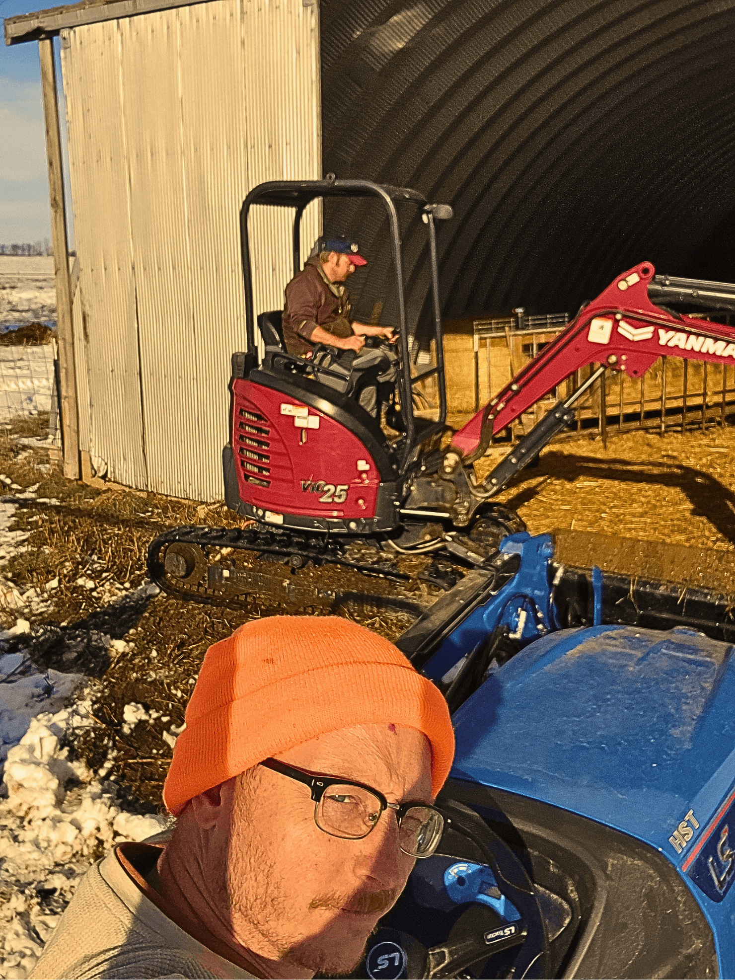 Man in orange beanie on blue tractor with a red mini excavator working nearby.
