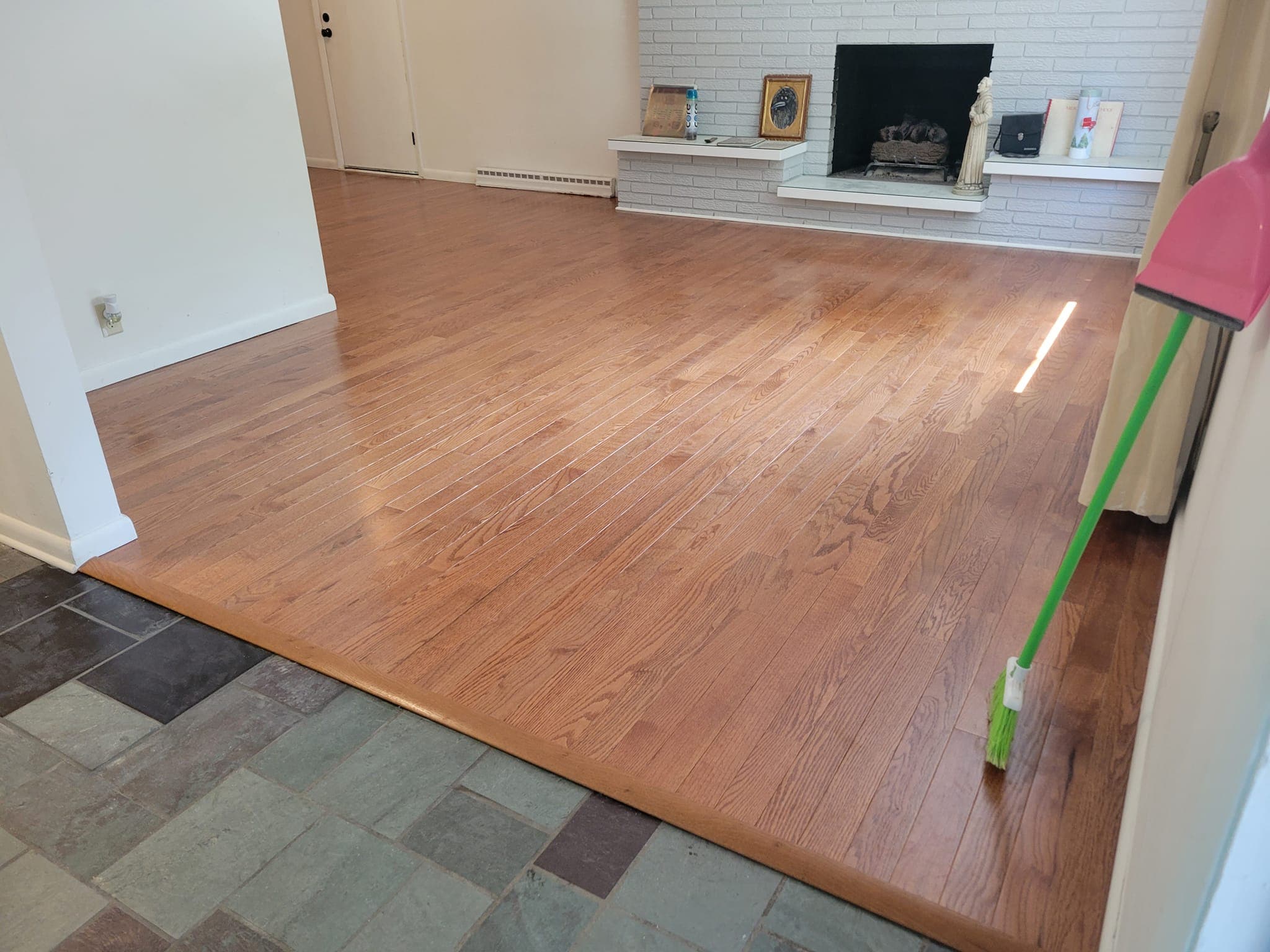 Polished light brown hardwood floor meeting grey slate tiles near a white brick fireplace.